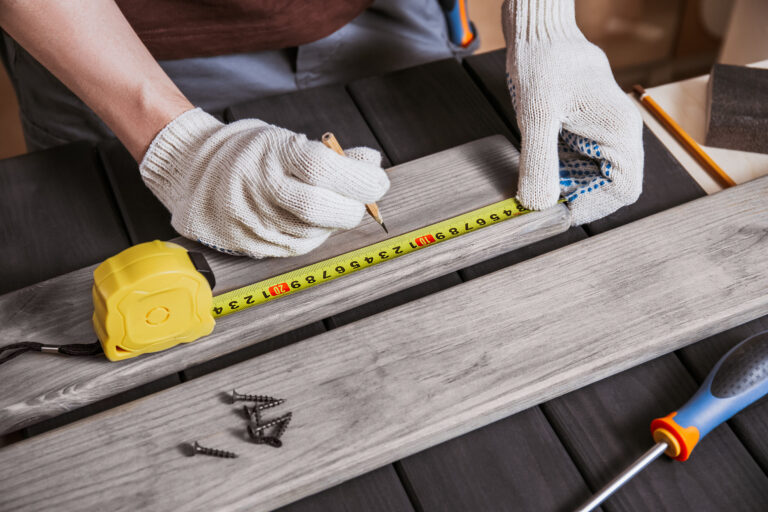 Hands in gloves of joiner in carpentry. He is successful entrepreneur at his workplace. Carpenter is measuring length of wood planks or timbers by measuring tape or ruler. Carpenter workspace.